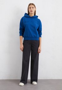Felpa blu con logo in rilievo, abbinata a pantaloni neri a gamba larga e sneakers bianche. Tessuto morbido al tatto, design casual.