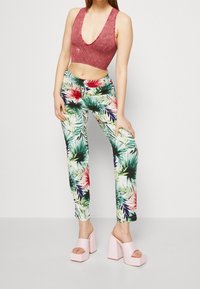 Femme portant un haut cropped sans manches à décolleté en V profond rose, un pantalon à imprimé tropical et des sandales à plateforme à talons roses clairs, posant devant un fond uni.
