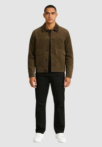 Giacca di velluto a coste marrone con tasche frontali, camicia nera sotto, pantaloni neri e sneaker bianche. Il modello è in piedi contro uno sfondo grigio.