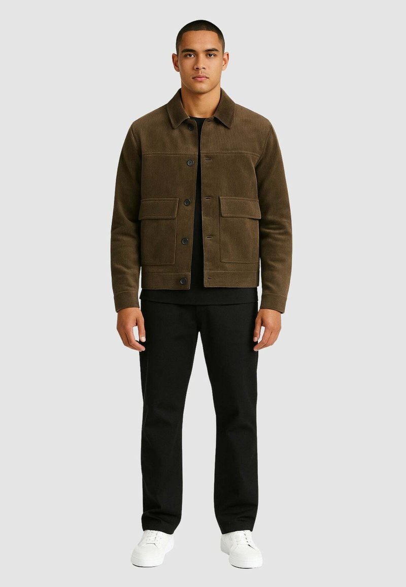 Giacca di velluto a coste marrone con tasche frontali, camicia nera sotto, pantaloni neri e sneaker bianche. Il modello è in piedi contro uno sfondo grigio.