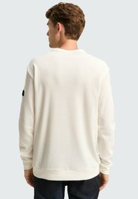 Witte langjarige shirt met een gestructureerde afwerking, ronde halslijn en geribbelde zoom. Bevat een zwart logo-oplaag op de linker mouw.