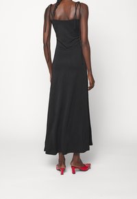 Czarna maxi sukienka z regulowanymi ramiączkami, dopasowanym korpusem i zwiewną spódnicą. Nosi ją w połączeniu z czerwonymi sandałami na obcasie, o gładkiej fakturze i minimalistycznym designie.