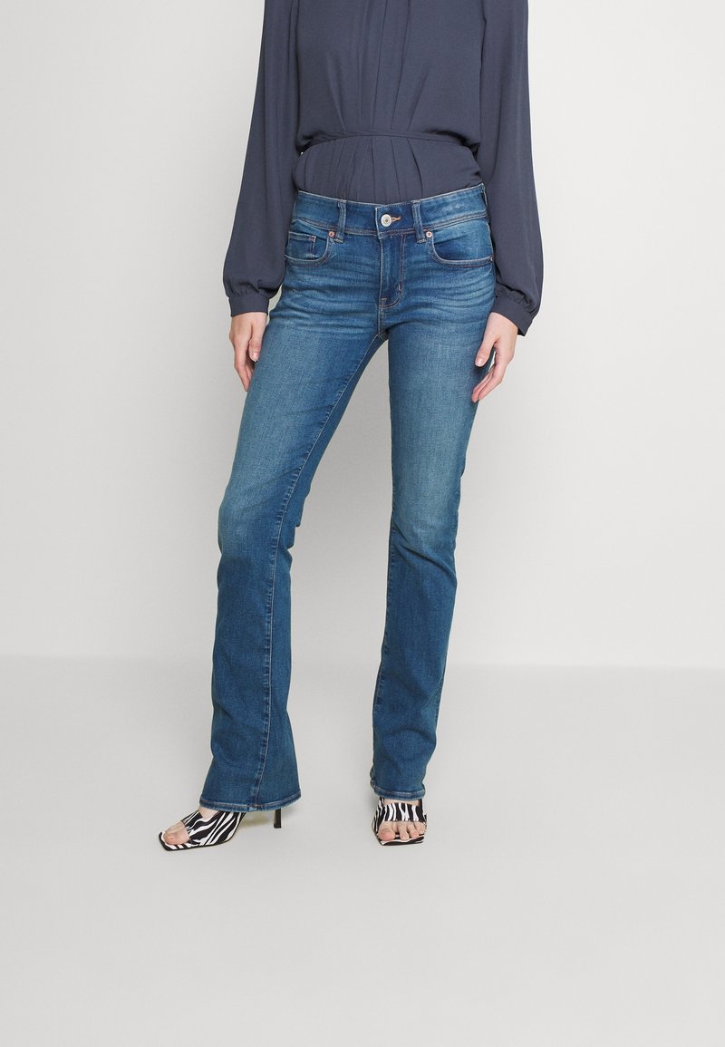 Kraftiga jeans i medelblått med hög midja, matchade med en mörk blus och svarta och vita randiga öppna klackar.