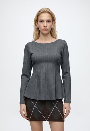 Jeune femme aux cheveux blonds ondulés portant un haut gris cintré à manches longues et une jupe marron à motif losange sur un fond blanc uni.