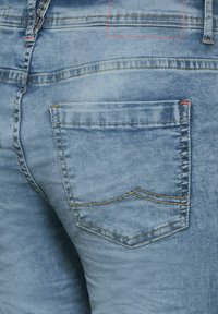 Light blue denim jeans featuring a back pocket with a curved top seam and red stitching accents. The fabric has a faded texture.