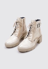 Botines beige con un acabado brillante y texturizado, que cuentan con un cierre de cordones en la parte delantera, cremallera lateral y un tacón cuadrado. Acentuados con una hebilla plateada.