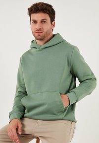 Vihreä puuvillainen huppari, jossa on etutasku, pitkät hihat ja kiristysnauhallinen huppu. Sileä rakenne ja rento istuvuus.