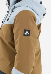 Ruskea ja vaaleansininen ulkoilutakki, jossa on tyylikäs muotoilu, logopatch ja säädettävä huppu, valmistettu kestävästä kankaasta.