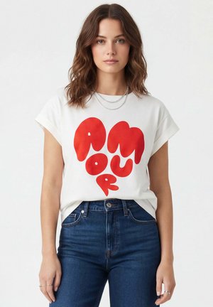 Femme aux cheveux bruns ondulés porte un t-shirt blanc avec un texte rouge audacieux « AMOUR » et un jean bleu taille haute sur fond uni.