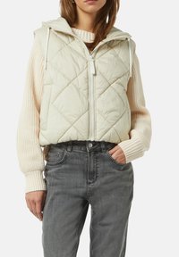 Kurtka pikowana w jasnym beżu z kapturem, z przodu na zamek błyskawiczny oraz białymi sznurkami. W połączeniu z kremowym swetrem dzianinowym i szarymi dżinsami.