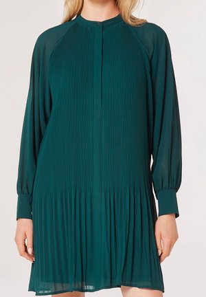 Femme portant une robe longue plissée vert foncé à manches longues avec col et poignets boutonnés, debout devant un fond uni.