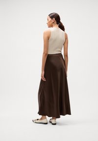 Top sans manches beige tricoté associé à une jupe maxi en satin fluide de couleur marron foncé. Le modèle porte des ballerines claires avec des semelles foncées.