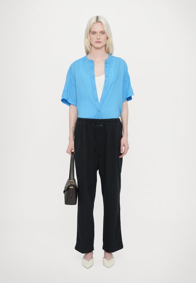 Camicia azzurra chiara a maniche corte con bottoni, abbinata a pantaloni neri larghi. Il modello tiene una piccola borsa scura e indossa scarpe a punta color crema.