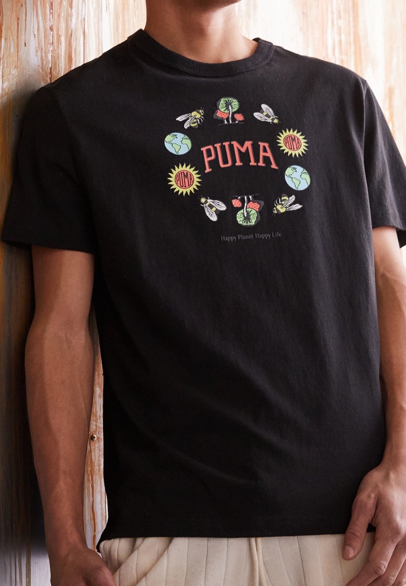 T-shirt noir avec le logo PUMA entouré d'illustrations d'abeilles, d'arbres, de la Terre et d'icônes du soleil, porté par une personne debout contre une surface en bois.