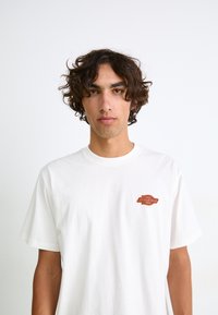 Hvid bomulds t-shirt med et lille orange og brunt logo på venstre bryst. Korte ærmer og en afslappet pasform giver komfort.
