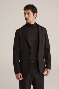 Homme portant un costume marron foncé avec une veste à deux boutons et un pull col roulé assorti, regardant sur le côté sur un fond uni.
