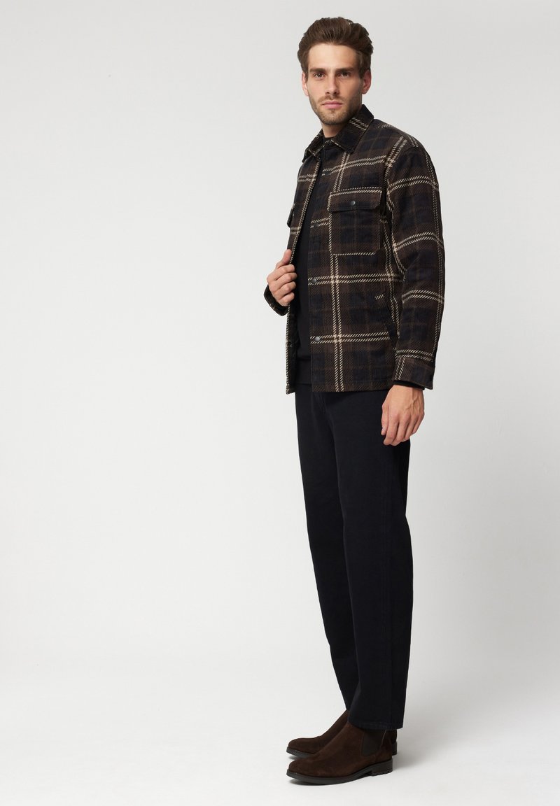 Koszula w kratę w ciemnych odcieniach z wzorem w kratę, z przednią kieszenią i zapięciem na guziki. Ciemne spodnie i brązowe botki za kostkę.