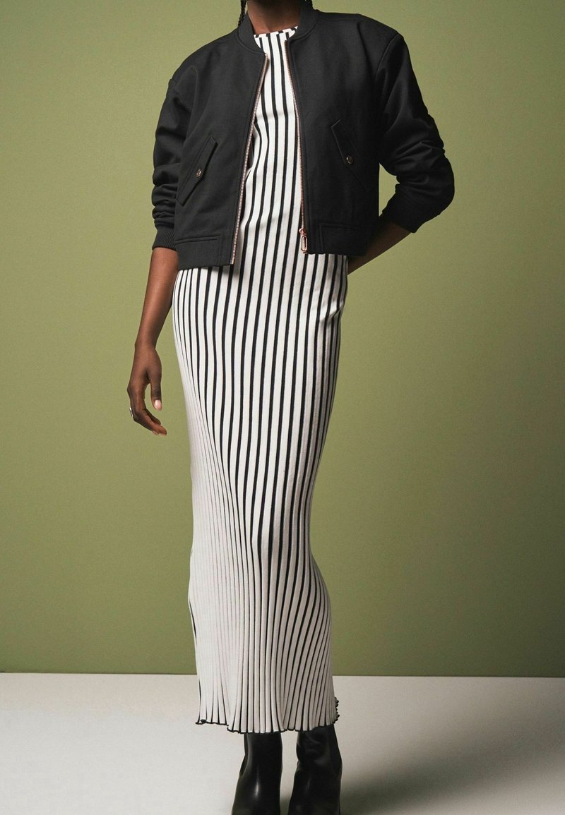 Chaqueta bomber negra sobre un vestido ajustado de rayas blancas y negras. El vestido tiene un diseño largo y fluido con un dobladillo ligeramente deshilachado.