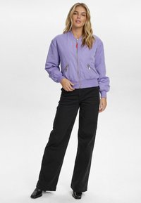 Purpura bomber jaka ar rāvējslēdzēja kabatām, ribotām piedurknēm un apakšmalu. Uzvilkta pār melniem platiem biksēm un melniem norādītiem apaviem. Minimalistisks dizains.