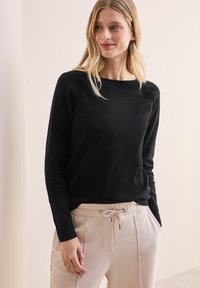 Schwarzer Langarmpullover mit Rundhalsausschnitt und Raglanärmeln, kombiniert mit hellen Jogginghosen mit Kordelzug. Glatte Textur.