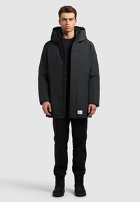 Czarna wodoodporna parka z kapturem, z zamkiem błyskawicznym, bocznymi kieszeniami i metką logo na dole, noszona na czarne spodnie i buty.
