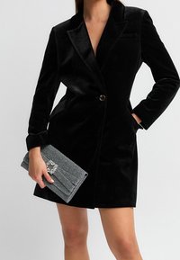 Schwarzer Samtblazer mit einem Ein-Knopf-Verschluss, Reverskragen und aufgesetzten Taschen, kombiniert mit einer silbernen, funkelnden Clutch mit einer dekorativen Brosche.