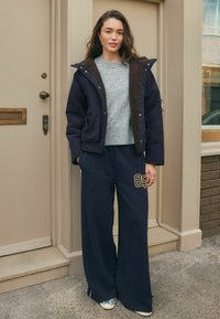Kvinna står utomhus iklädd en marinblå jacka med huva och brun foder, grå tröja, marinblå byxor med vida ben och en "85"-patch, samt silverfärgade sneakers.