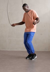 Man in peach t-shirt and blue pants skipping rope indoors on gray floor and wall.