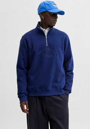 Persoon in een marineblauwe sweatshirt met een kwart ritssluiting en de tekst "ORIGINALS Studio", met een heldere blauwe pet, die opzij kijkt.