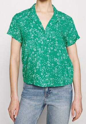 Zielona bluzka z krótkim rękawem w biały kwiatowy wzór, noszona na wierzchu jasnoniebieskich dżinsów.