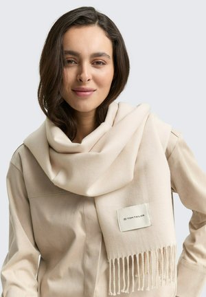 Écharpe en laine beige avec des bords frangés et une étiquette. Drapée sur l'épaule, elle présente une texture douce et un design minimaliste.