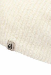 Soft, cream-colored ribbed knit fabric with a small beige label featuring a black crowned emblem sewn on the edge.