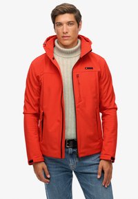 Veste softshell rouge avec capuche, équipée d'une poche zippée, accents noirs, et un pull crème texturé en dessous. Portée avec un jean bleu.