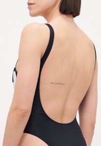 Maillot de bain une pièce noir avec des bretelles larges, un dos découpé et une texture lisse. Comprend un accent blanc et un tatouage dans le dos.