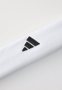Hvid sport håndledsband lavet af tekstureret stof med et sort broderet logo med tre striber, skrå design og ren finish.