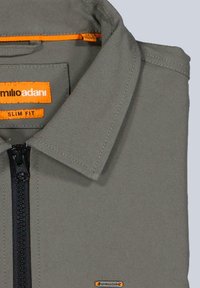 Graues Slim-Fit-Hemd mit einem schwarzen Reißverschluss, Kragen und kontrastierenden orangenen Akzenten auf dem Etikett und dem Öffnungsbereich. Glattes, strukturiertes Material.