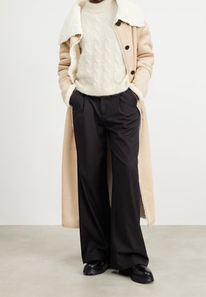 Personne portant un long manteau beige en peau lainée, un pull tricoté torsadé blanc, un pantalon noir à jambes larges et des chaussures noires épaisses, debout les mains dans les poches.