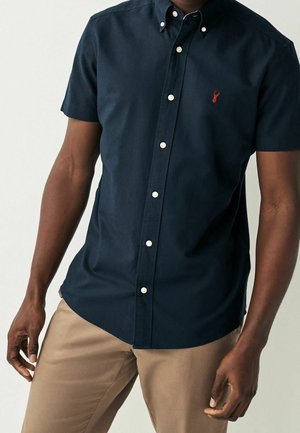Homme portant une chemise bleu marine à manches courtes avec des boutons blancs et un logo de cerf rouge, associée à un pantalon beige.