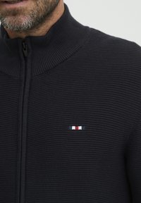 Tengerészeti zipzáros dzseki bordás textúrával, magas gallérral és egy kis hímzett logóval, amely piros, fehér és kék csíkokkal díszíti a mellet.