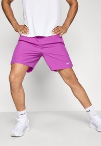 Homme en chaussures de sport blanches, chaussettes blanches, chemise blanche et shorts de sport roses vifs, se tenant avec les jambes écartées et les mains sur les hanches.