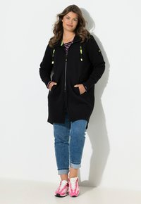 Chaqueta negra con capucha y cordones de neón verde, tela texturizada, cremallera frontal, bolsillos laterales, combinada con vaqueros de mezclilla y zapatillas deportivas rosas.