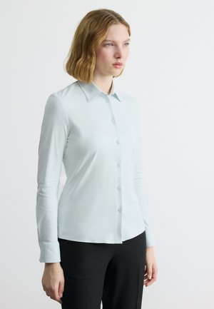 Žena s blond vlasy po ramena, nosící světle modrou košili s knoflíky a černé kalhoty, stojící před bílým pozadím.