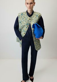 Geprinte vest in groen en goud met blauwe accenten, gedragen over een donker shirt en broek. Het model houdt een felblauwe, gestructureerde tas vast.