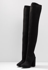 Botas negras hasta la rodilla, hechas de ante suave, con tacón cuadrado y punta afilada, con un diseño sin costuras y sin elementos de metal visibles.