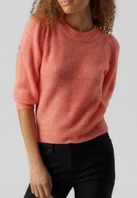 Koraalroze gebreide trui met een ronde hals, driekwartmouwen en ribbelstructuur, gedragen met zwarte broek.