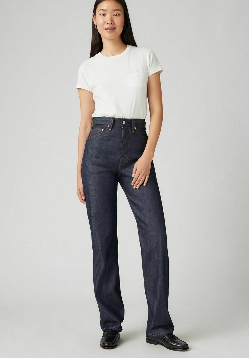 Jeansy z wysokim stanem, w ciemnoniebieskim kolorze, z prostą nogawką, delikatnymi przetarciami i pomarańczowymi przeszyciami, połączone z dopasowaną białą koszulką.