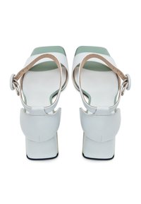 Une paire de sandales à talons blocs ouvertes blanches en cuir, avec des brides autour de la cheville et des semelles vertes, vues de l'arrière sur un fond blanc.