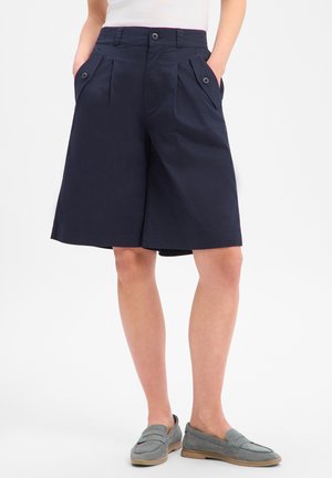 Donna che indossa pantaloncini blu navy fino al ginocchio con pieghe davanti e tasche laterali con bottoni, abbinati a mocassini grigi slip-on.