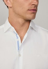 Chemise en coton blanc avec un col pointu, petits détails de rayures bleues le long de la patte de boutonnage et un logo brodé discret sur la poitrine.