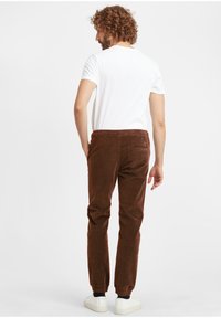 Brown corduroy pants with an elastic waistband, tapered fit, and side pockets, paired with a white T-shirt and white sneakers.